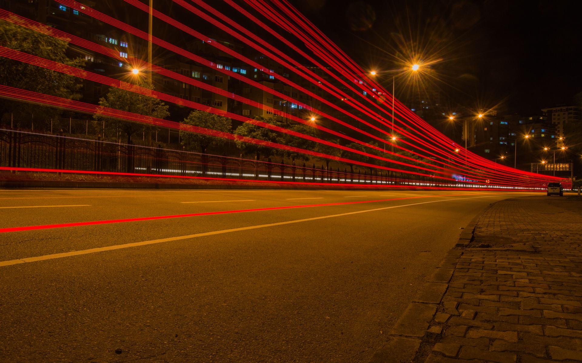 A long exposure of an empty city road at night, red tail-light trails sweeping past under amber streetlights.