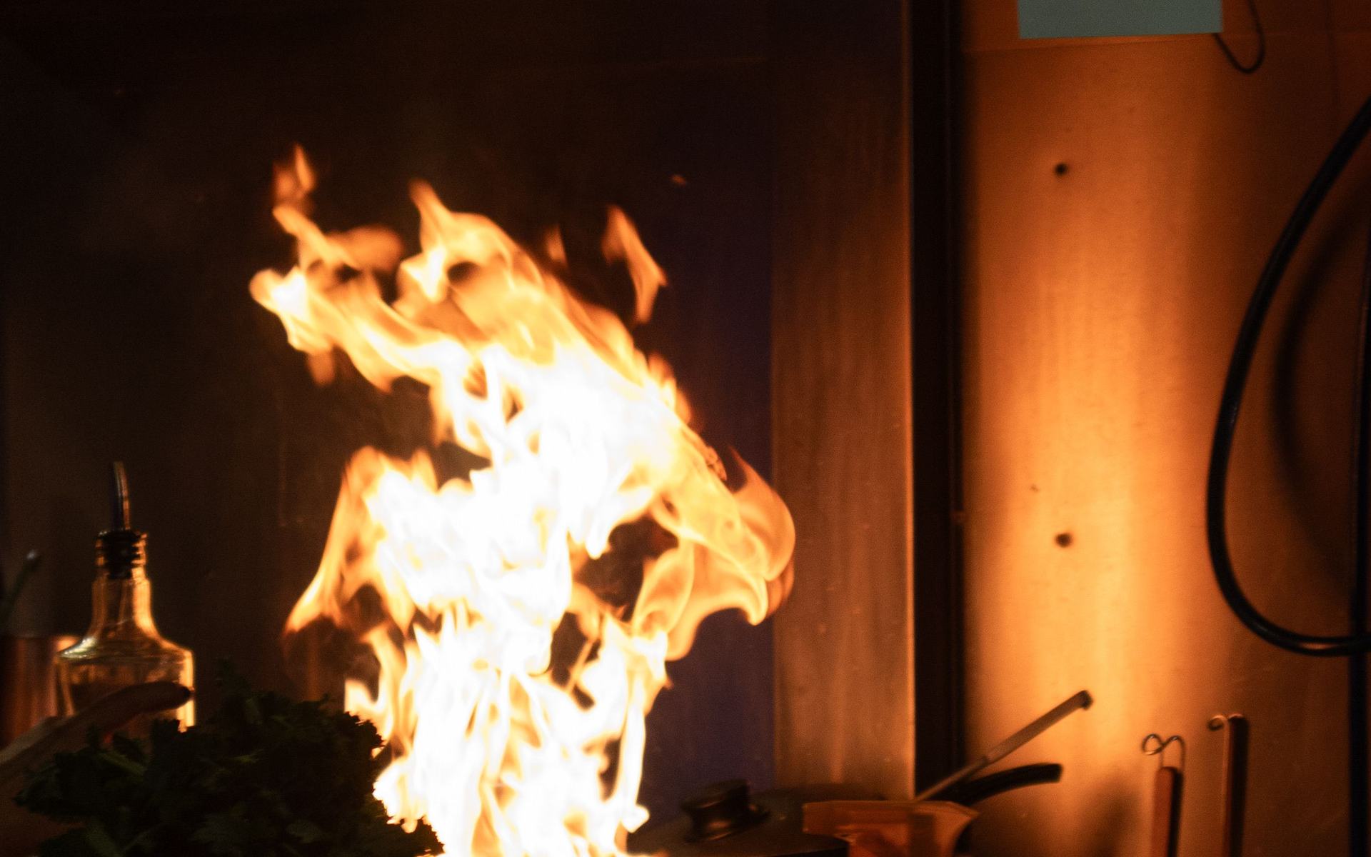 A tall flame leaping from a pan in a restaurant kitchen, lighting a copper wall.