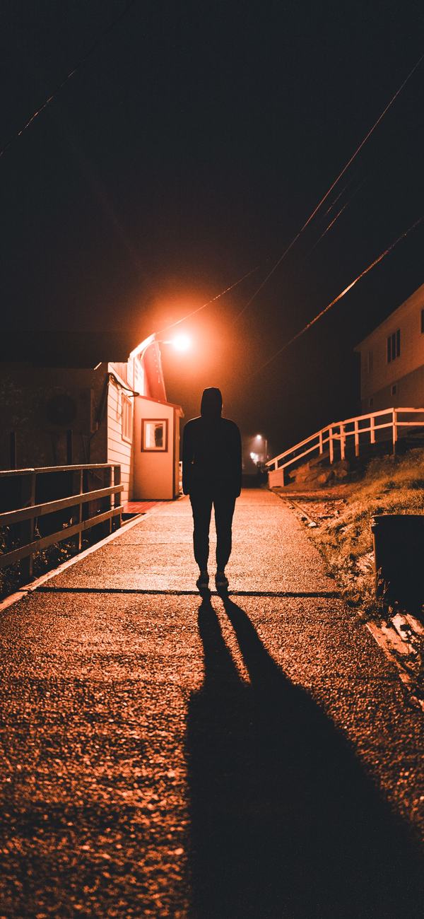 A hooded figure, seen from behind, on a wet path at night under a single orange streetlamp.