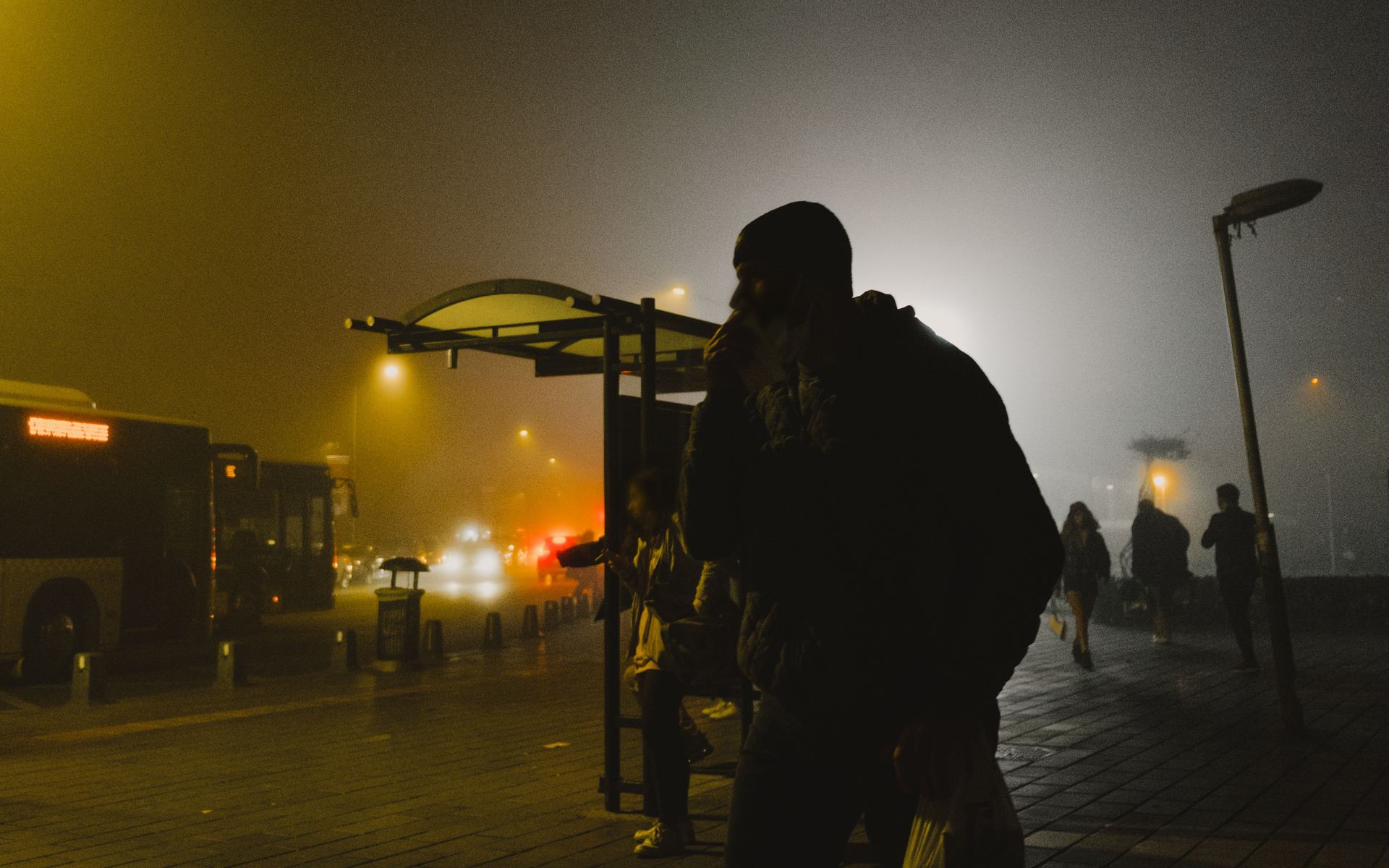 Silhouetted commuters at a bus shelter on a foggy night, lit by amber streetlights.